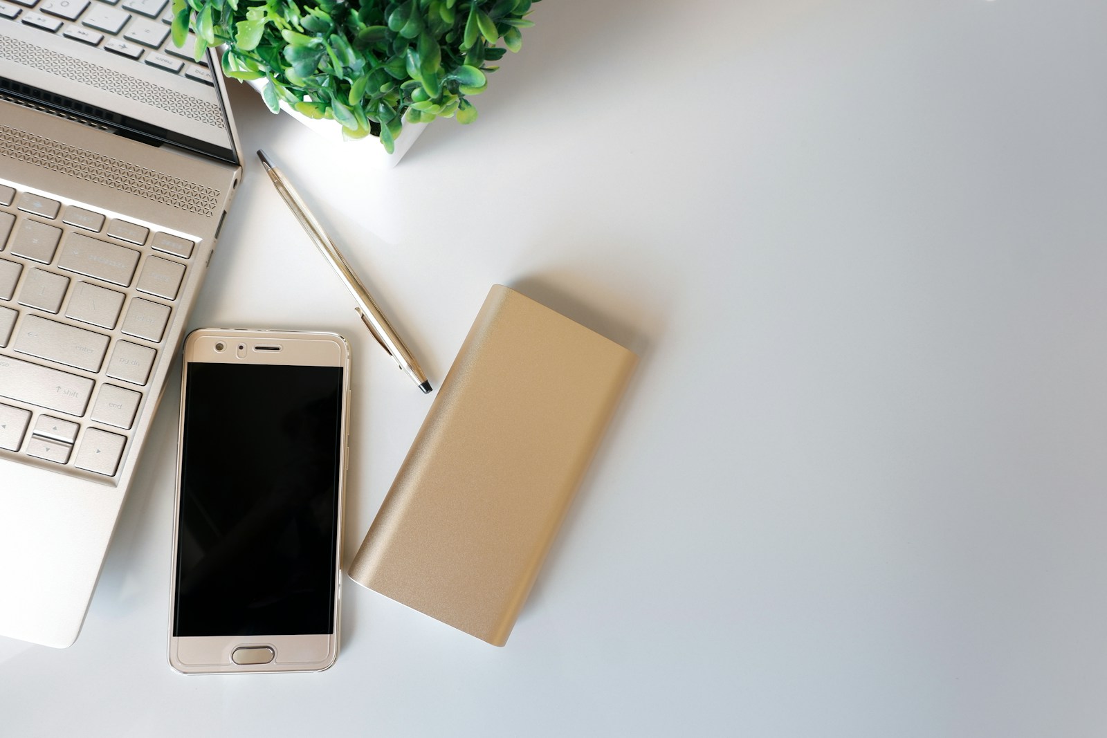 Laptop, smartphone, pen, gold power bank and small green plant on a white desk.